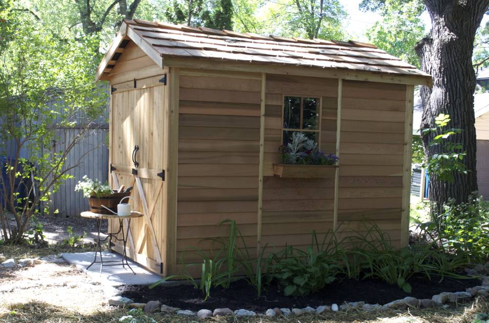 A new wooden shed with a flower bed around the side ringed by rocks.