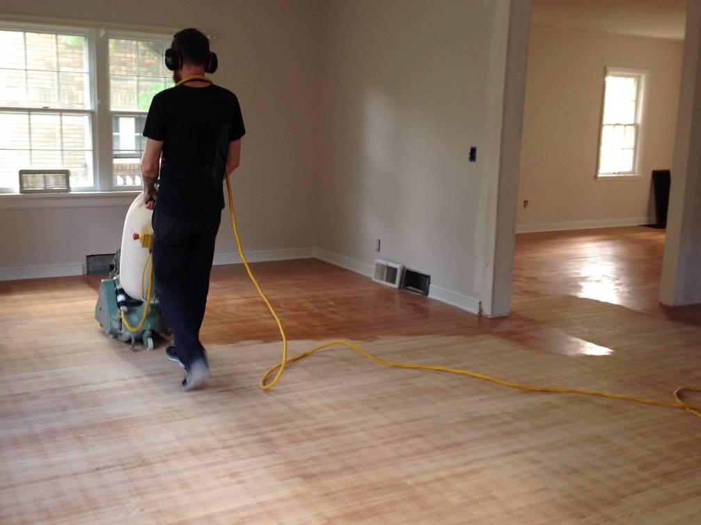A man with ear muffs is pushing an electric floor waxer across a wooden floor.