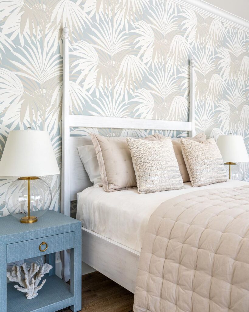 White palm leaves on walls with light blue and pink colors in wallpaper behind white bed with blue end table.
