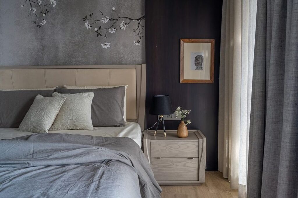 Grey wallpaper with japanese flowers and bed with grey comforter and beige end table.